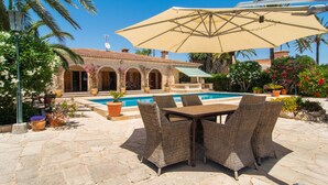 Ein Poolbereich mit Tisch und Stühlen, einem großen Sonnenschirm und einem Gebäude mit Bögen im Hintergrund.