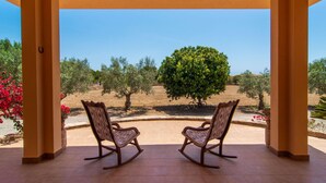 Eine überdachte Terrasse mit zwei Schaukelstühlen, die auf eine Olivenhainlandschaft und einen klaren Himmel blicken.