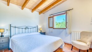 A bedroom with a bed, bedside tables, a chair, a window with a view, and wooden ceiling beams. - Amazing home in Cala Ratjada (Cala Ratjada)