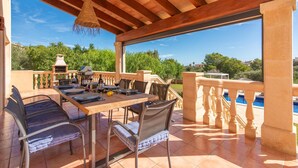 Area pranzo all'aperto con tavolo in legno apparecchiato per quattro persone, circondato da sedie, una lampada a sospensione e vista su piscina e vegetazione.