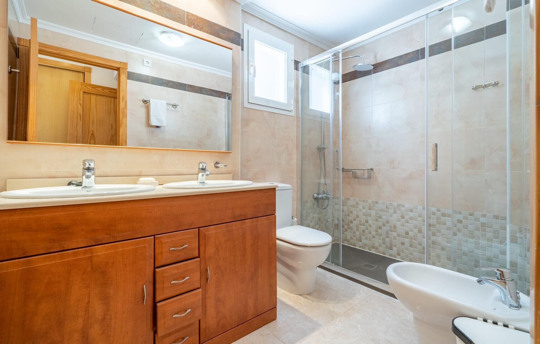 A modern bathroom with a wooden vanity, two sinks, a toilet, and a glass-enclosed shower area.