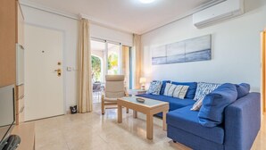 Una sala de estar moderna con un sofá azul, una mesita y una silla. Se ve una puerta que da a un espacio exterior con una mesa y sillas.