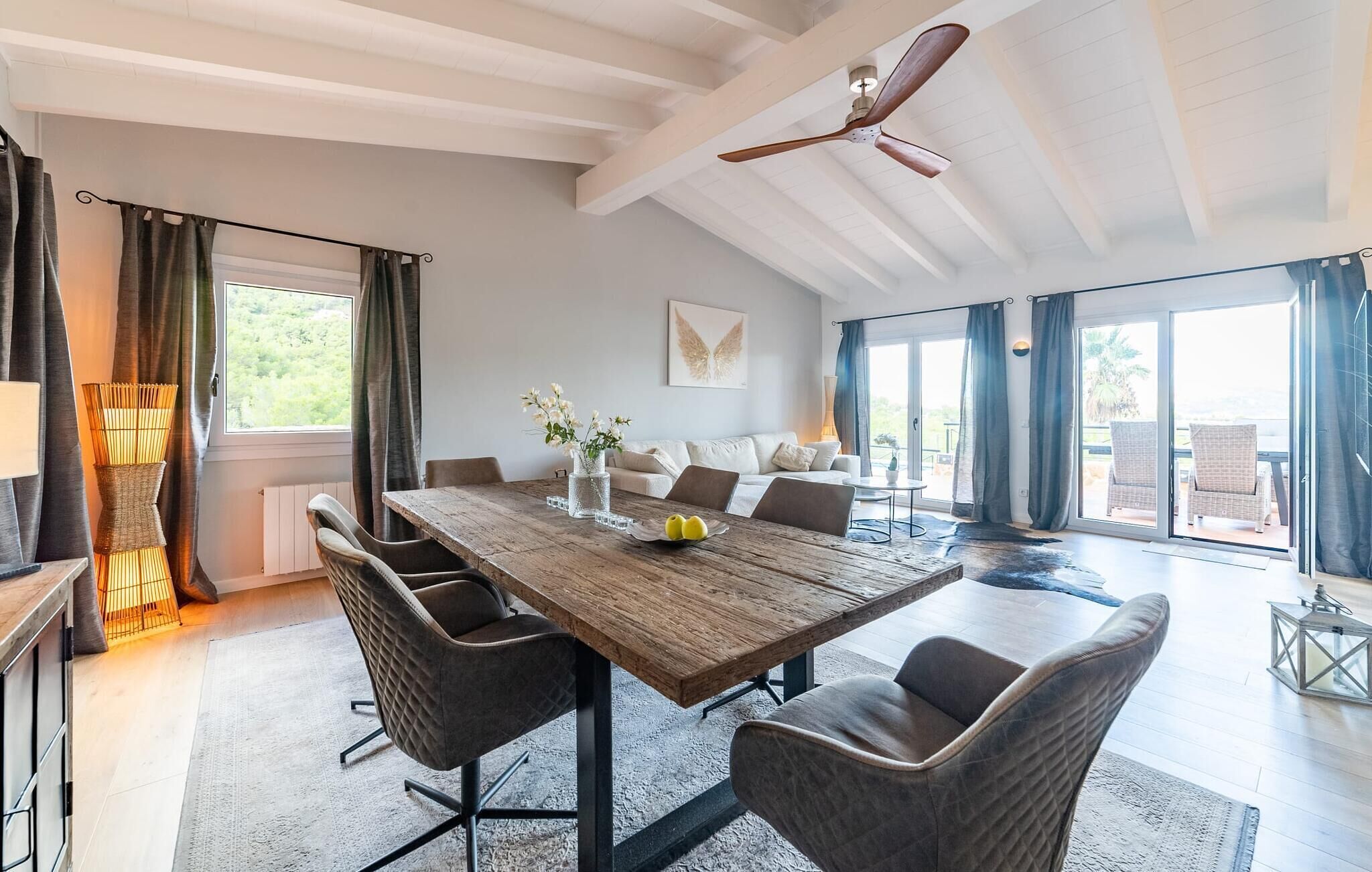 A spacious living room with a large wooden dining table, comfortable seating, a ceiling fan, and large windows offering a view of the outdoors.
