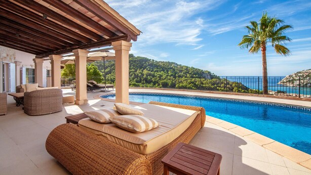 A poolside area with a wicker sofa and a wooden table, overlooking a clear blue pool and a mountainous landscape. - Lovely home in Port d'Andratx with sauna (Port d'Andratx)