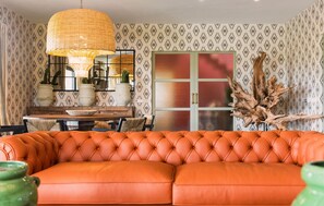 Una habitación con un sofá naranja con capitonado, una mesa de comedor con sillas, una lámpara colgante y papel tapiz estampado.
