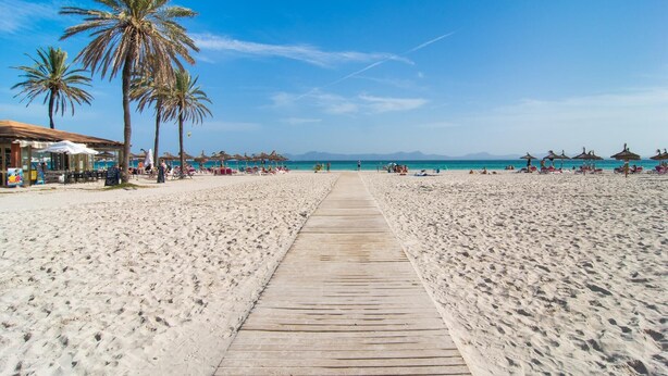 A beach with a wooden boardwalk leading to the sea, palm trees, beach umbrellas, and a few people. - 3 bedroom gorgeous home in Alcudia (Alcudia)