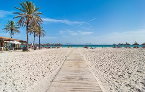 Ein Strand mit einer Holzpromenade, die zum Meer führt, Palmen, Strandstühlen und einigen Personen.