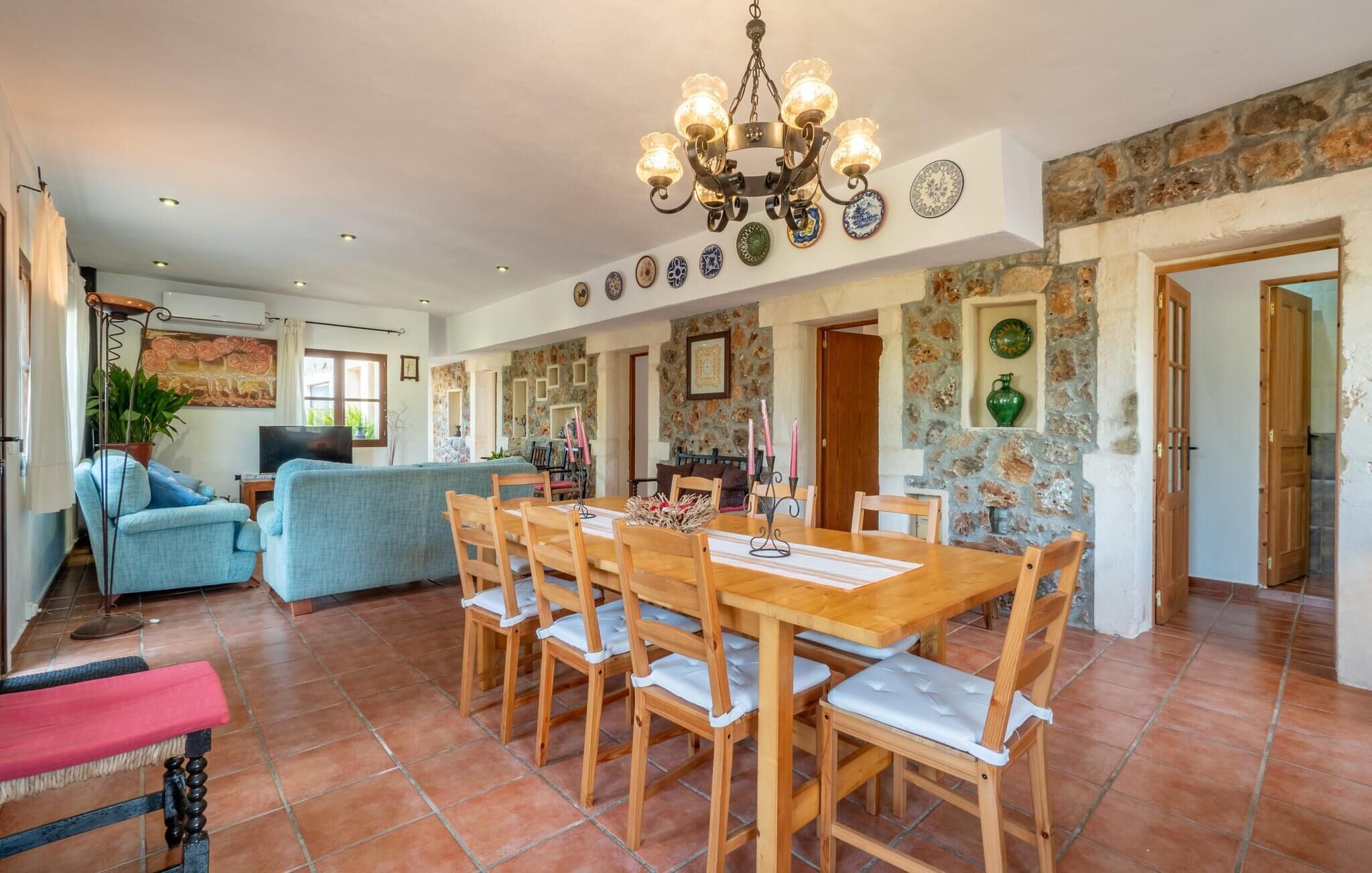 A spacious dining area with a wooden table and chairs, a chandelier, and a stone wall with decorative plates.