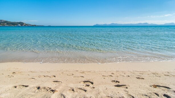 Ein Strand mit klarem Wasser, Fußspuren und Bergen im Hintergrund.