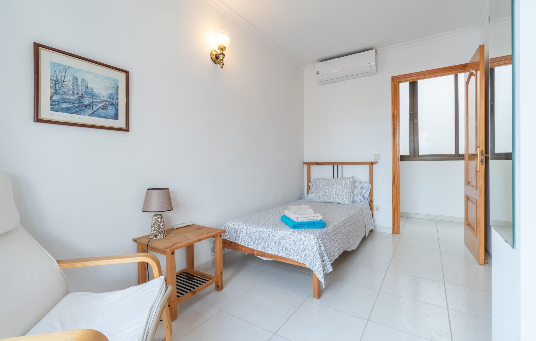 A small, clean room with a bed, a chair, a table, and a painting on the wall.