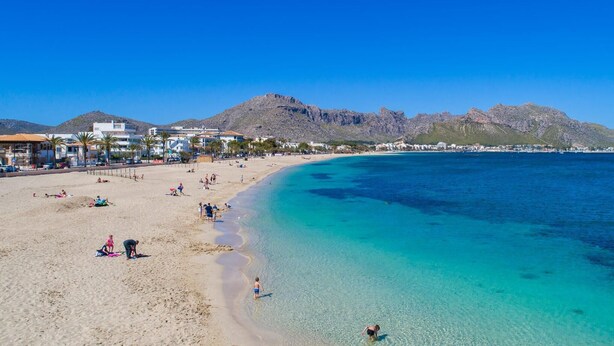 Une plage aux eaux turquoise et limpides, un rivage sablonneux et des bâtiments visibles au loin.