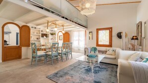 Ampla sala de estar com mesa de jantar, cadeiras, sofá e mesa de centro de vidro. Escada leva a um andar superior, e o espaço apresenta design moderno com vigas de madeira e lustre.