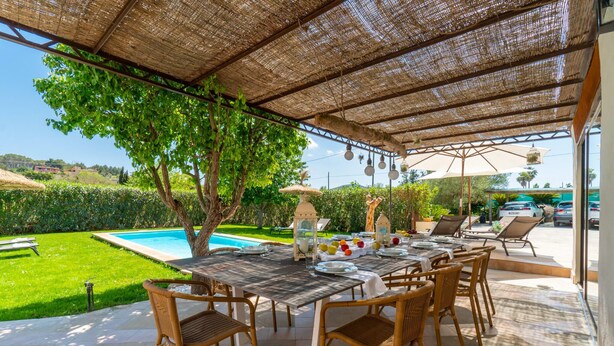 Um pátio externo com uma mesa posta para refeições, rodeado por cadeiras de vime, piscina e vista para um jardim exuberante.