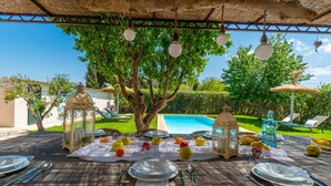 Uma área de refeições ao ar livre com mesas preparadas para uma refeição, cercada por vegetação exuberante e uma piscina ao fundo.