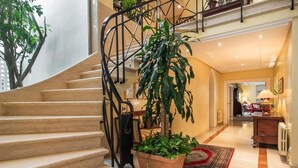 A staircase with a black metal railing leading to a second floor. There are potted plants on the landing and a patterned rug on the floor.