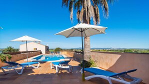 Un área de piscina en la azotea con sillas de descanso y sombrillas, rodeada por un muro bajo y palmeras.