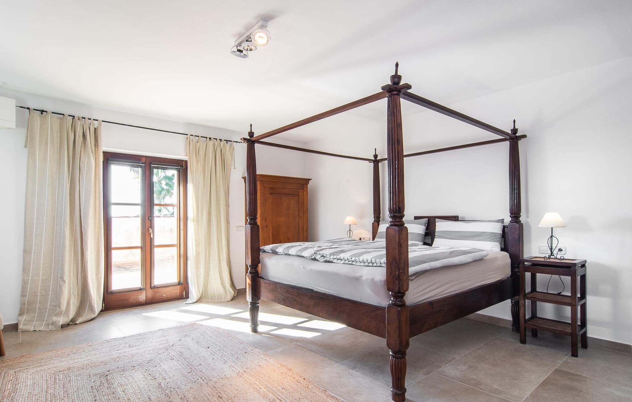 A four-poster bed in a well-lit room with a wooden wardrobe, a small bedside table, and a door leading to an outdoor area.