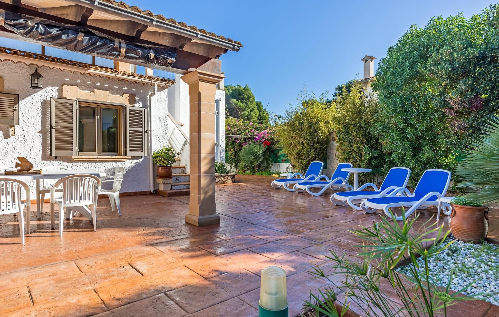 Un patio con mesa y sillas, una zona de estar cubierta con sillones azules, y un jardín con diversas plantas y árboles.