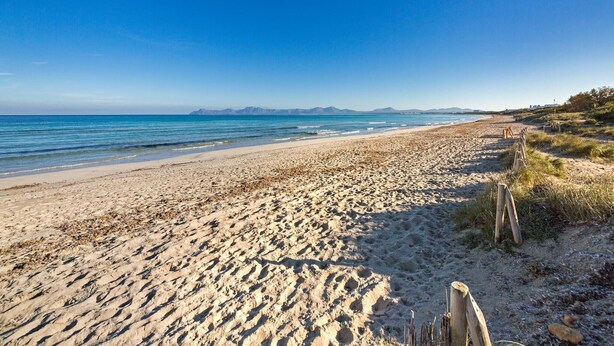 Ein weiter Sandstrand mit sanften Wellen, blauem Himmel und Bergen in der Ferne.