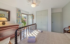 Une chambre à coucher comprenant un lit, une commode, un ventilateur de plafond, une fenêtre avec des rideaux et une porte.