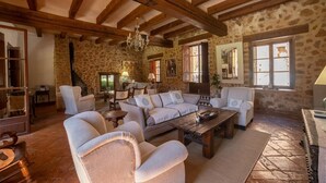 Een rustiek interieur met zichtbare houten balken, stenen muren, een open haard en een gezellige zithoek met een bank en fauteuils.