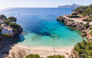 Una playa costera con aguas turquesas cristalinas, acantilados rocosos y algunos edificios.