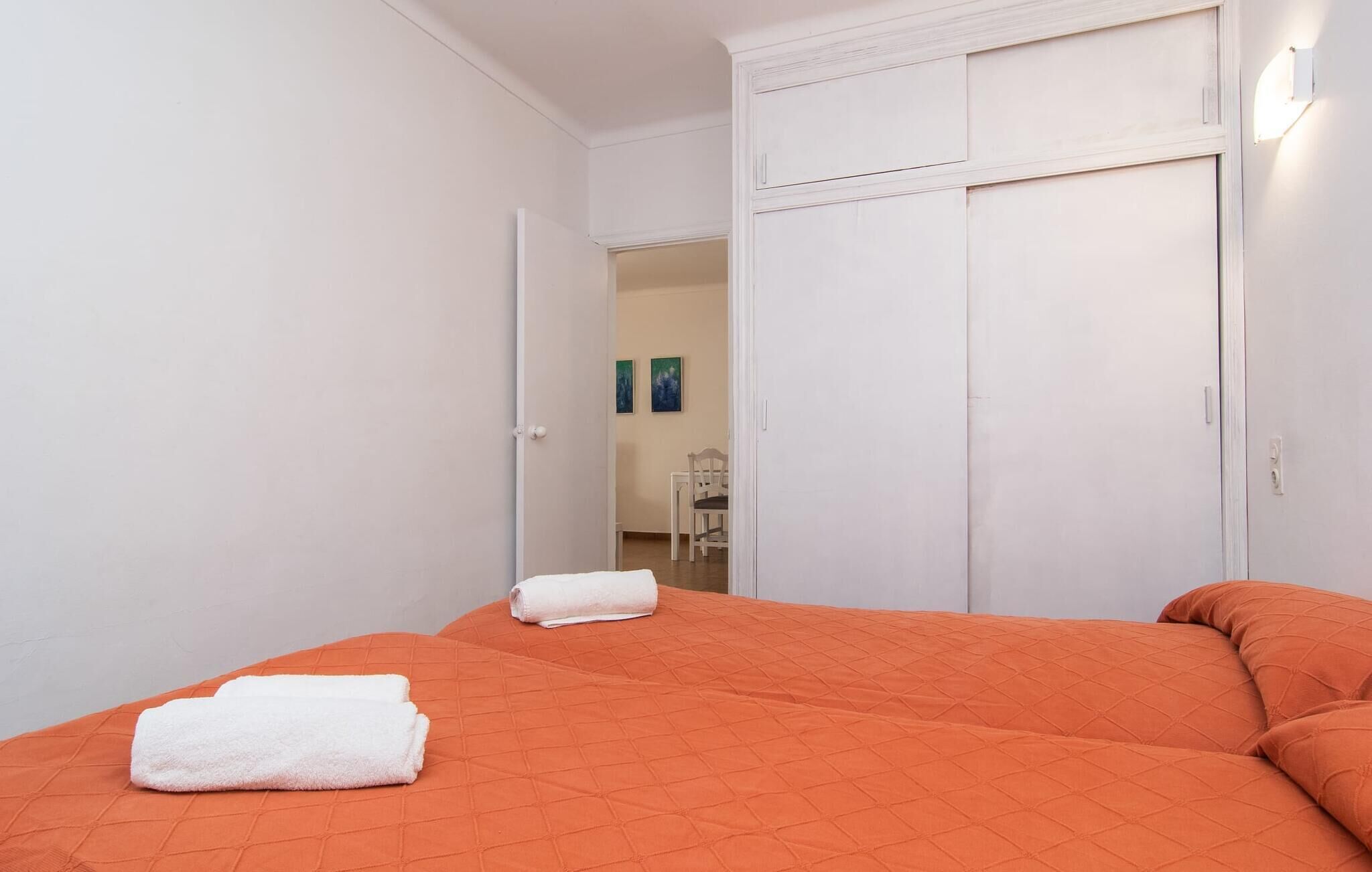A hotel room with two beds, each with an orange quilted bedspread and white towels. There is a white wardrobe with a mirror and a door leading to a bathroom.