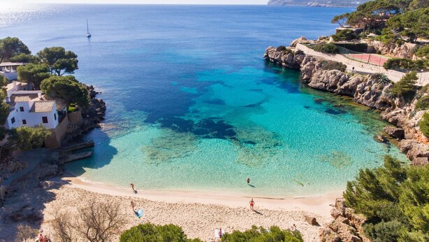 Ein Küstenstrand mit klarem türkisblauem Wasser, steinigen Klippen und einigen Gebäuden.