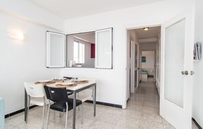A compact hotel room with a dining area featuring a table set for two, a black chair, and a white chair. The room has a white door, a small hallway leading to another room, and a wall-mounted mirror. - Gorgeous apartment in Cala Ratjada with outdoor swimming pool (Cala Ratjada)
