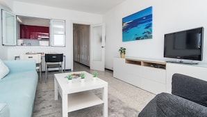 Ein modernes Wohnzimmer mit einem weißen Sofa, einem grauen Sofa, einem Couchtisch, einem Fernseher, einem Strandbild und einer Küchenzeile mit Schränken und einer roten Arbeitsplatte.