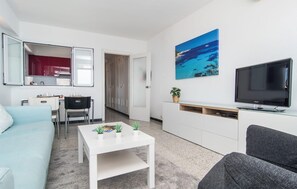 A modern living room with a white sofa, a grey sofa, a coffee table, a TV, a painting of a beach, and a kitchen area with cabinets and a red countertop. - Gorgeous apartment in Cala Ratjada with outdoor swimming pool (Cala Ratjada)