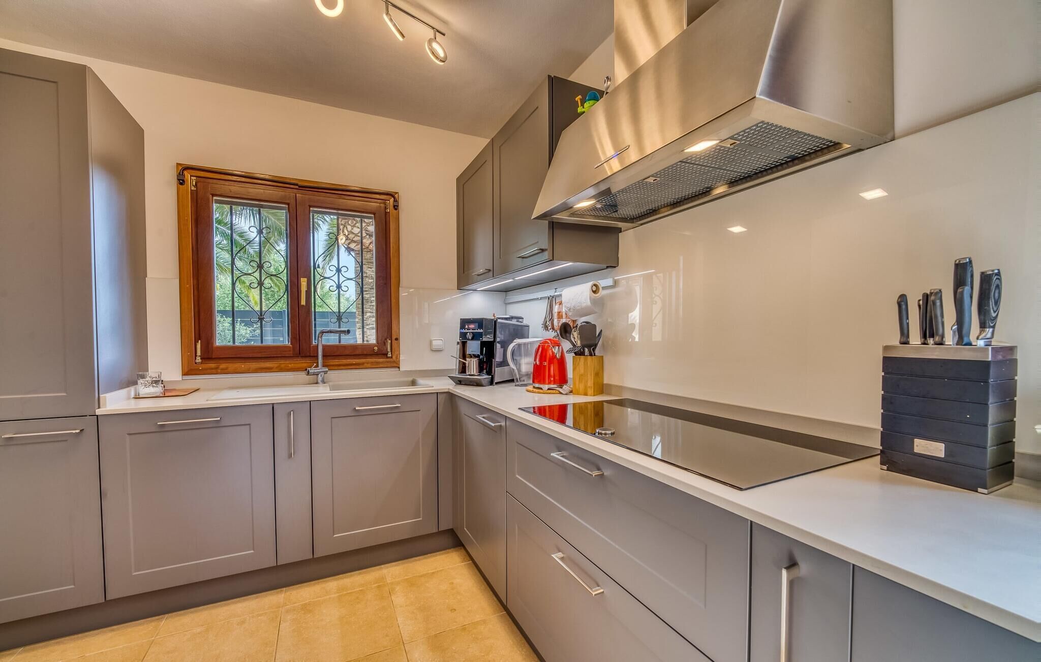 A modern kitchen with grey cabinets, a stainless steel range hood, a built-in oven, and a window with decorative iron bars.
