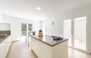 A modern kitchen with a central island, white cabinetry, and a large window providing natural light. - Beautiful home in Sa Font de Sa Cala (Sa Font de Sa Cala)