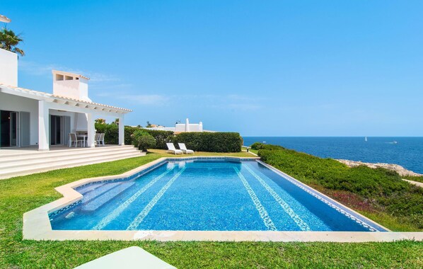 A swimming pool with a clear view of the ocean, a white building with a covered patio, and a well-maintained lawn. - Beautiful home in Cala dOr with private swimming pool, can be inside or outside (Cala dOr)