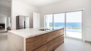Una cucina moderna con un piano di lavoro in marmo, mobili in legno e vista sull'oceano attraverso ampie porte scorrevoli.