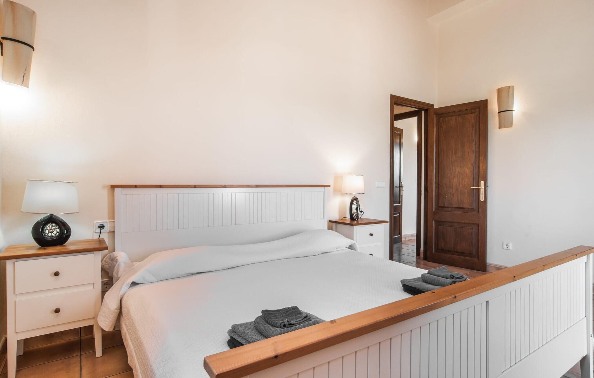 Une chambre à coucher avec une tête de lit en bois, un couvre-lit blanc, deux tables de chevet avec des lampes, une petite table de chevet en bois et une porte en bois.