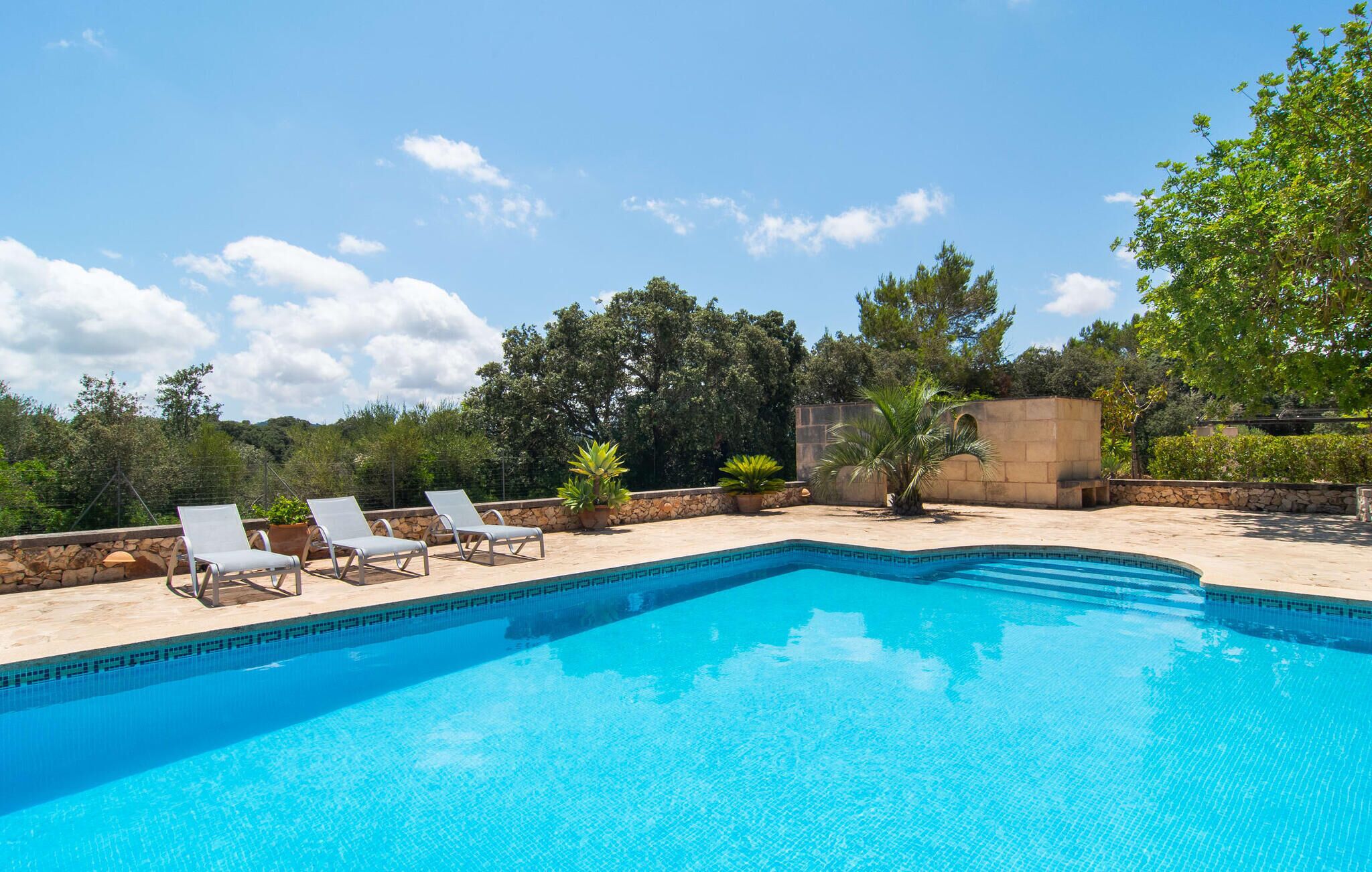 A rectangular swimming pool with a tiled edge, surrounded by a stone deck. There are three lounge chairs aligned parallel to the pool's edge.