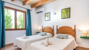 Two single beds with white linens and towels, a window with blue curtains, an air conditioner, and a framed leaf artwork on the wall. - Beautiful home in Alcudia with private swimming pool, can be inside or outside (Alcudia)