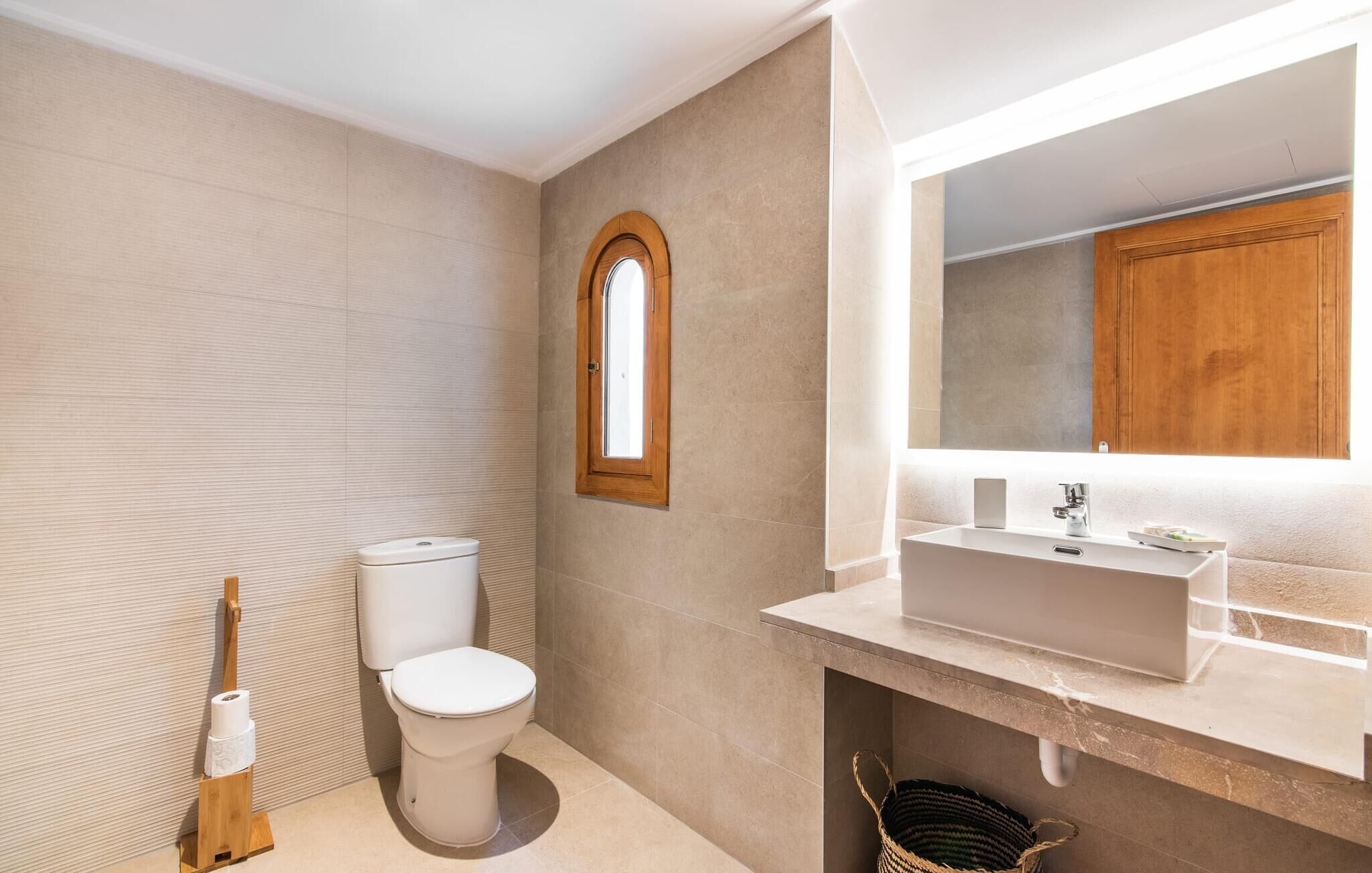 A bathroom with a toilet, a wooden toilet brush, a sink with a mirror, and a window.