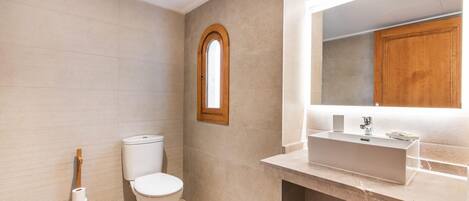 A bathroom with a toilet, a wooden toilet brush, a sink with a mirror, and a window.