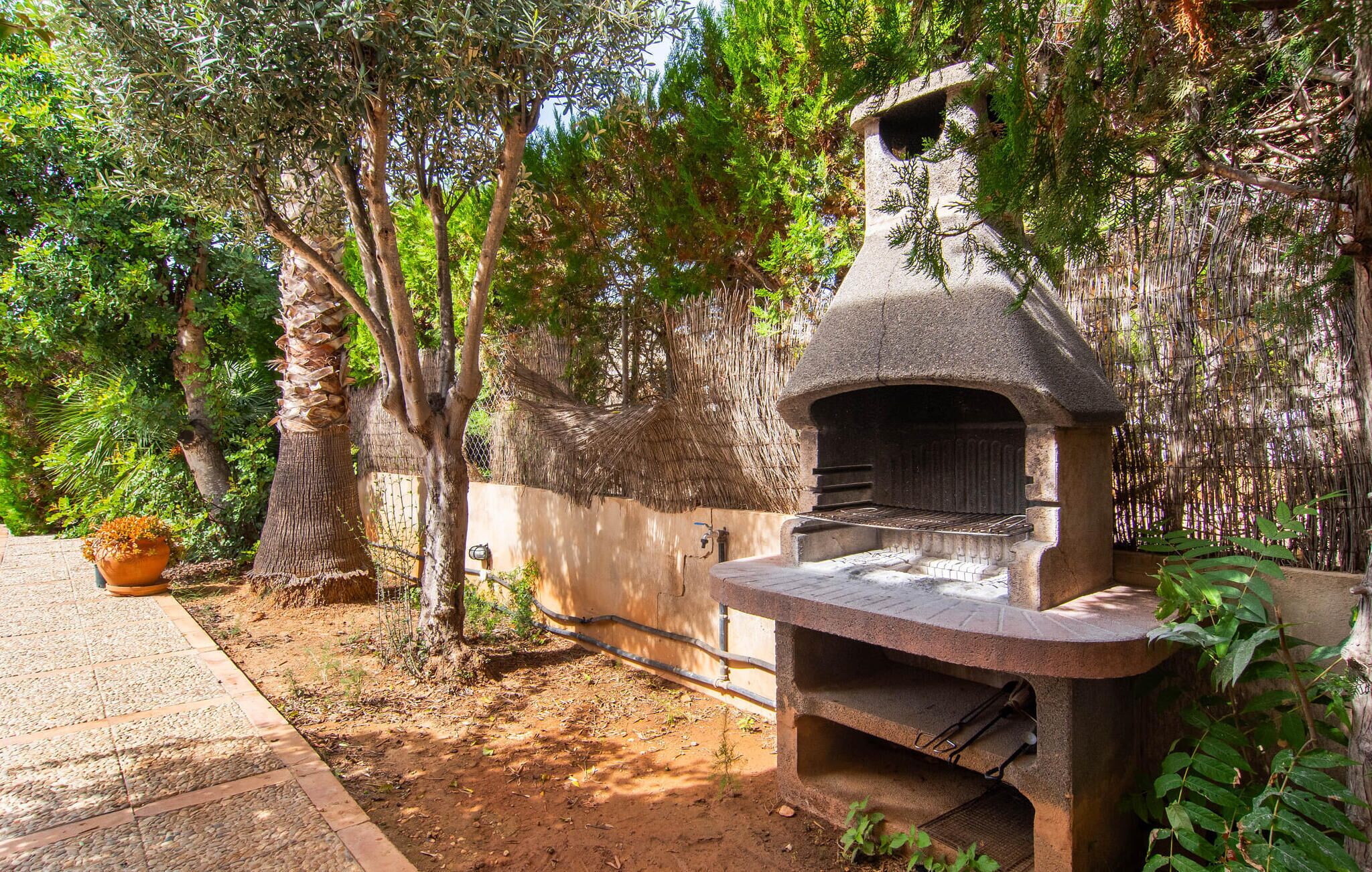 Un four à pizza en pierre, installé à l’extérieur, dans un jardin agrémenté d’arbres et d’une plante en pot à proximité.