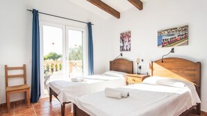 Ein Hotelzimmer mit zwei Betten, einem Holzsessel, einem Balkon mit blauen Vorhängen und einem Bild einer Zugszene an der Wand.