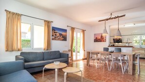 Ein modernes Wohnzimmer mit Sofa, Couchtisch und Essbereich mit Stühlen. Zwei Fenster mit Vorhängen, ein Gemälde an der Wand und eine durchgehende Küchenzeile.