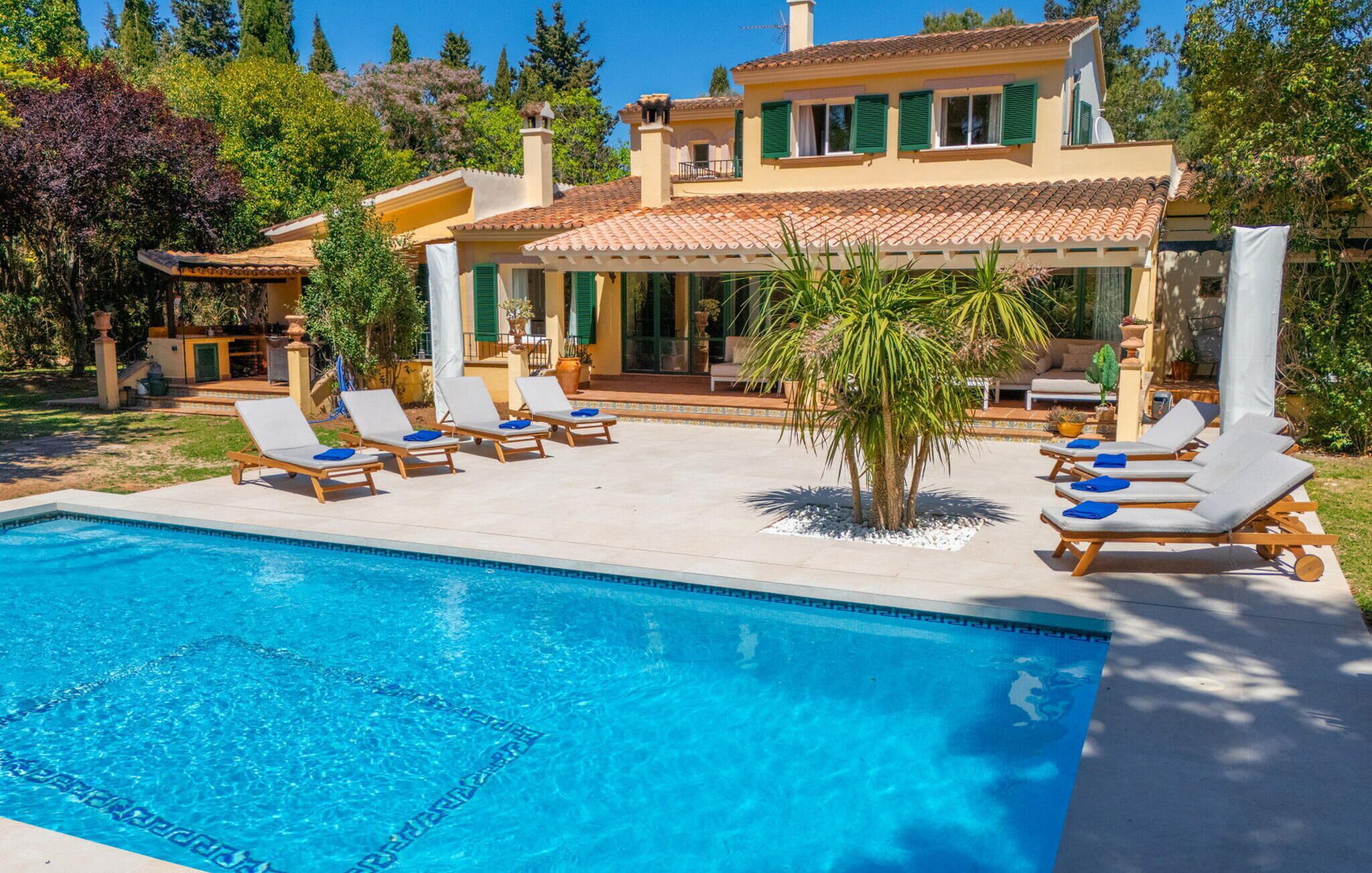 A pool with lounge chairs, a patio area, and a house with a tiled roof and green shutters.