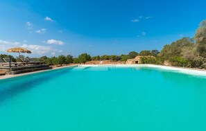 Ein großer, klarer blauer Pool mit Liegestühlen und Sonnenschirmen, umgeben von Bäumen und einem Steinhaus.
