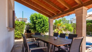 Eine Terrasse im Freien mit einem Holztisch und Stühlen, umgeben von Grünpflanzen und einem Schwimmbecken im Hintergrund.