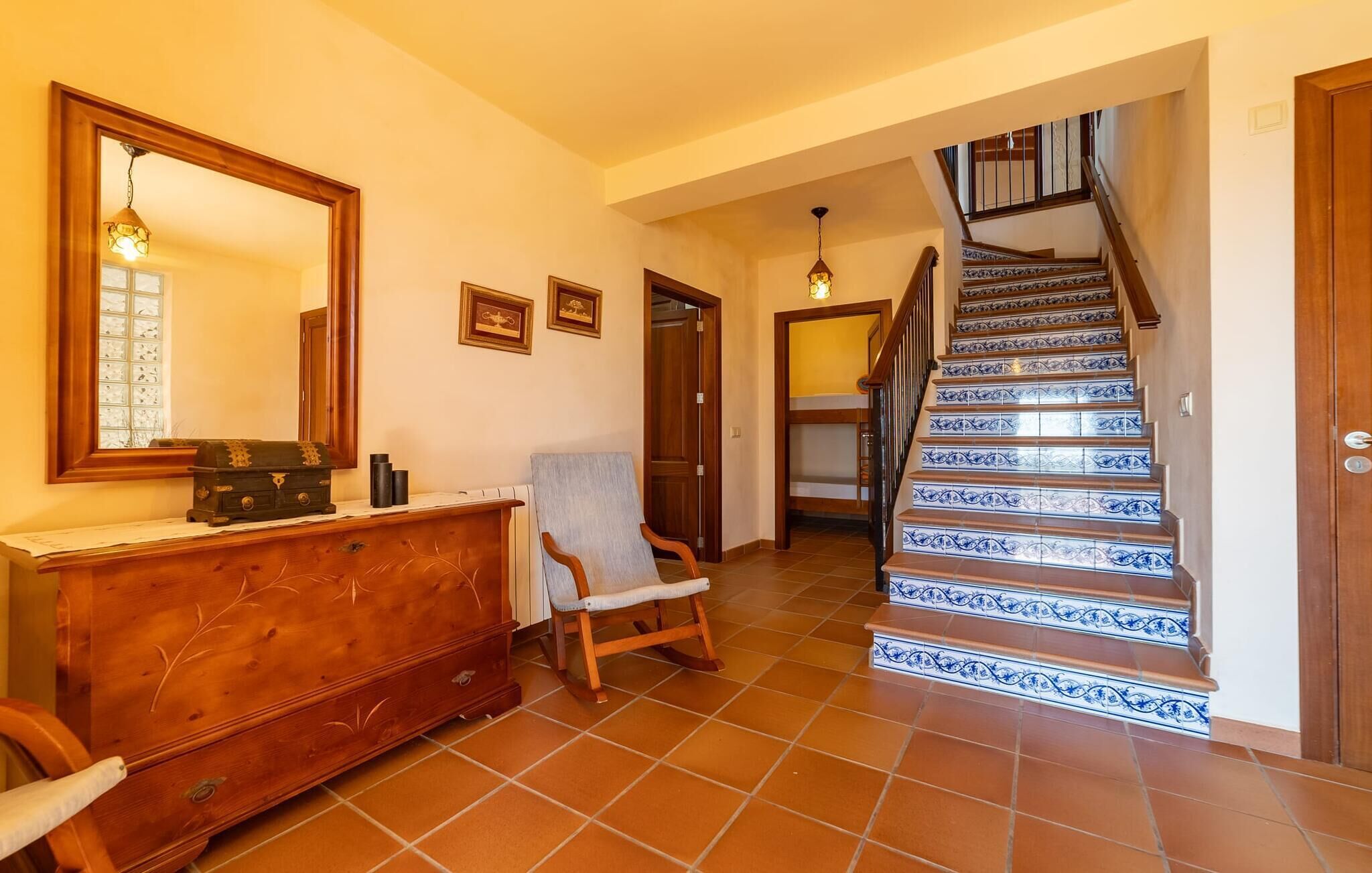 A hallway with a wooden dresser, a rocking chair, a staircase with blue and white tiles, and a mirror on the wall.