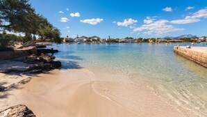 Kystlandskab med klart vand, en sandstrand og bygninger i baggrunden.