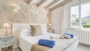 A bedroom with a stone wall, a large bed with white bedding, two bedside lamps, and a view of greenery outside. - Lovely home in Pollenca with private swimming pool, can be inside or outside (Pollenca)