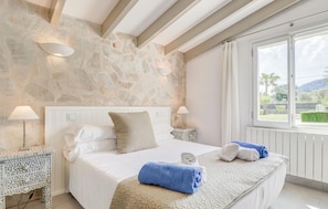 A bedroom with a stone wall, a large bed with white bedding, two bedside lamps, and a view of greenery outside. - Lovely home in Pollenca with private swimming pool, can be inside or outside (Pollenca)